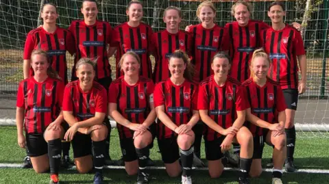 A women's football team pose for a photo in two rows. Some kneel on the front row whilst others stand behind them. All wear red and black strips.