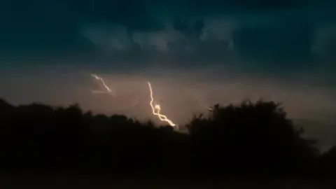Carolyn R/Weather Watchers Lightning in the middle of an orangey, grey sky over the silhouette of trees.