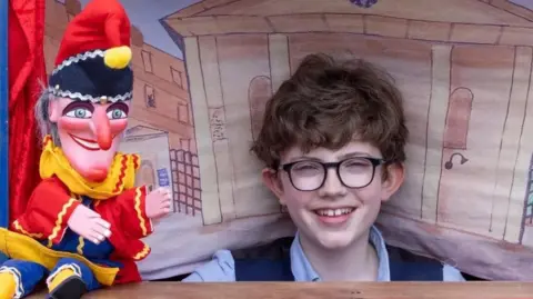 A smiling William Vaughan holding a Mr Punch puppet inside a performing tent.