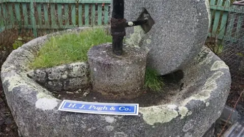 HJ Pugh and Co A stone mill with a stone tub and an upright wheel above it and a metal axle.