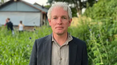 Danny Kruger looks at the camera - he is wearing a dark linen jacket and grey shirt. Blurred behind him is lots of greenery
