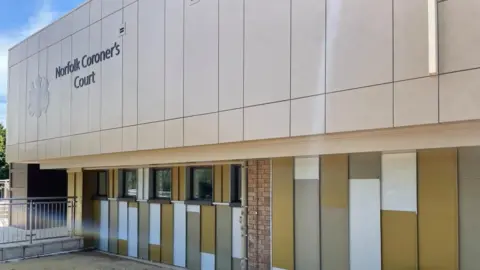 Andrew Turner/BBC Norfolk Coroner's Court is a gold-panel clad building, square in form, with white and mustard panels on the lower floor, with brown framed windows. The Royal crest adorns the building with signage stating: "Norfolk Coroner's Court".