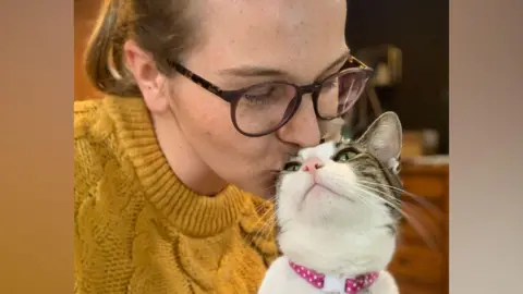A woman wearing brown glasses and a yellow knitted jumper kisses the head of a brown and white cat that has its head slightly raised toward her. It is wearing a pink collar with white polka dots on it. 