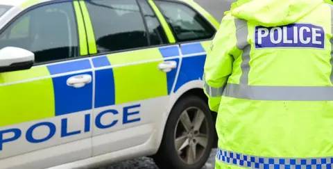 The picture shows the back of a police officer dressed in a yellow high-vis coat and a parked white, yellow and blue police car. On the back of the coat is the word "POLICE" in white letters on a rectangular blue background.