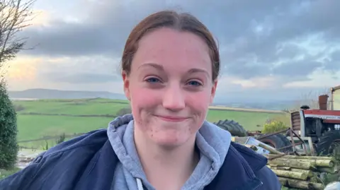 Jessie Young stands at her family farm, her brown hair is tied up and she is wearing a grey hoodie with a blue jacket. 