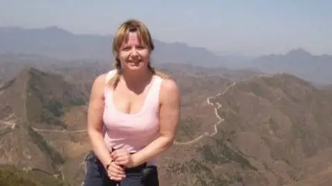 Kerr Family Dawn stands on a rocky viewpoint holding a walking pole, positioned in the foreground of a mountainous landscape. Rolling hills and winding paths stretch into the distance under a clear sky, creating a wide, elevated outdoor scene.