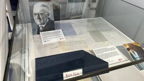 A glass display box containing a collection of letters and pictures. A picture at the top left shows a black and white picture of an older man in a suit and glasses. One at the bottom right shows a woman in a coat wearing a large scarf.