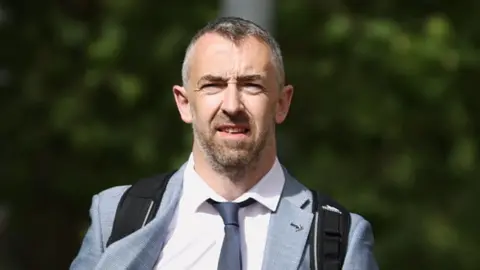 PA Media Daithí McKay pictured walking to court, he is wearing a grey suit with a white shirt and a grey tie. He has short grey hair and a beard. He is also wearing a black backpack.