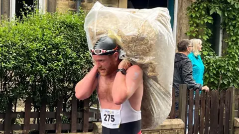 BBC/Dave Edwards A man runs with a bale of hay over his shoulder. He wears a black swimming cap and goggles. Two women, with their backs turned, watch from behind a wooden gate.