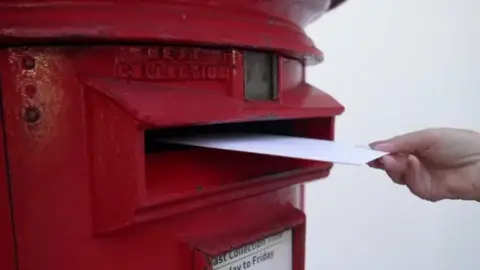 A hand posting a white envelope through a red post box opening. 