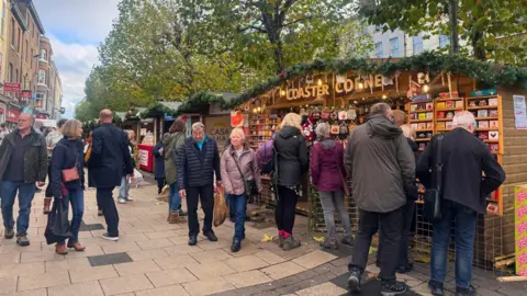 BBC/Emily Johnson A busy outdoor Christmas market with decorated wooden stalls and shoppers.