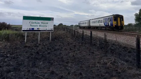A train is passing close to a burned field with a sign reading 'Coatham Marsh Nature Reserve' standing in it.