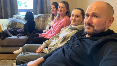 BBC Lachlan is sitting on a sofa with his two daughters and his wife Leeann. The family's dogs have couch all to themselves.