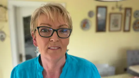 BBC A woman stands in her kitchen. She has blonde hair, glasses and is wearing a blue shirt.