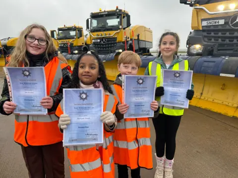 Four children holding certificates, standing in front of a row of snowploughs
