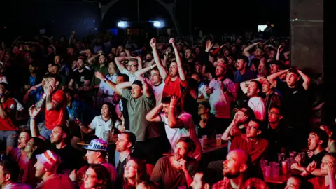 Alastair Brookes A large crowd of England football fans at a watch party in Bristol. Men and women can be seen and many are wearing football tops and other merch like hats. Many of the fans have their arms up in the air or on their head with their mouths open. 