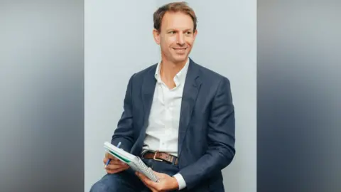 British Orthodontic Society Matthew Clover is wearing a white shirt, open at the neck, and a navy jacket and jeans. He is sitting down and has a pen and notepad in his hands.