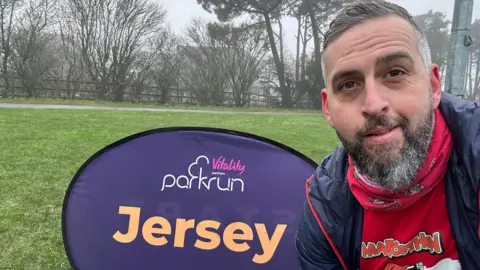 A selfie by Sam Wilkes, who is smiling at the camera. He is by a sign saying "Parkrun Jersey" on a field and he is wearing a dark blue coat with a red Marcothon running top underneath and a Marcothon neck scarf. He has short grey hair and beard.