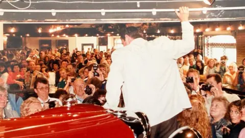 Jersey Pearl John Nettles is facing away from the camera, weaing a white blazer. He’s stood next to a red car and is hanging on to the roof of a room. He is surrounded by people watching him, many with cameras. 