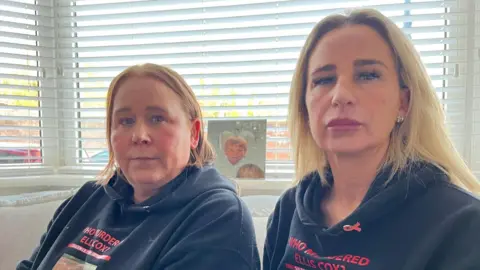 Carolyn Cox has long blond hair and is wearing diamond cross earrings. She is sat alongside Julie O'Toole who has mid length red hair. Both are sat on a living room sofa in a bay window with white vertical blinds. Behind them is a photograph of box in a heart shaped silver photo frame.