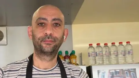 A man in a black and white striped T-shirt and a navy apron stands in a food truck. Behind him are lots of bottles of water and a transparent fridge with soft drinks in.