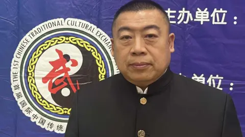 Picture features a man in black traditional Chinese martial arts clothing, standing in front of a blue wall with a logo featuring Chinese writing behind him. There is white writing is featured behind him.