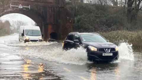 A car and a van drive through a deep puddle under a bridge, spraying water.