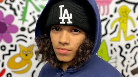 Kobie pictured in a black LA hat and a blue hoodie jacket. He has shoulder-length curly dark hair. He is standing behind a wall with colourful cartoons printed on it.