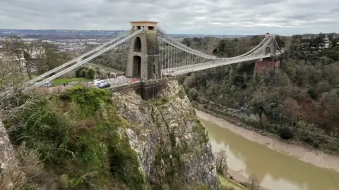 The Clifton Suspension Bridge is seen from a higher point in the Avon Gorge on a dry but cloudy day. The river is between high and low tide below the bridge and the North Somerset countryside is visible in the distance