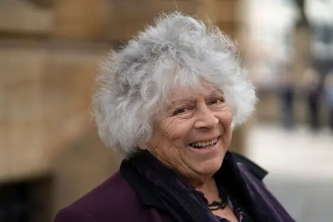Getty Images Miriam Margolyes smiling at the camera. She has white-grey hair sticking up. She is older and is wearing a purple jacket. She is wearing a black necklace across her chest. The background behind her is blurred.