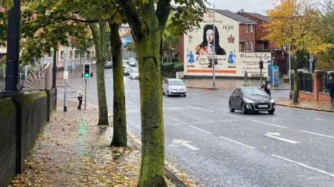 Scene showing a road with cars travelling across. There are autumn leaves on the ground and trees either side of the road. To the right, a mural to King William III.