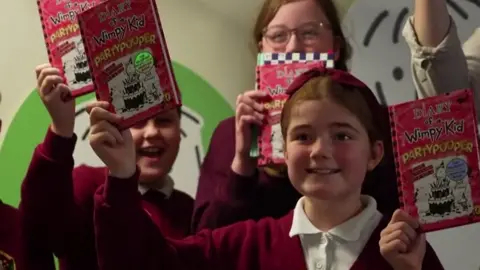 Three smiling children holding up Kinney's books. They are wearing maroon school uniforms.