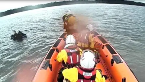An orange lifeboat with four crew members wearing white helmets and red life jackets. There is a mattress on the water.