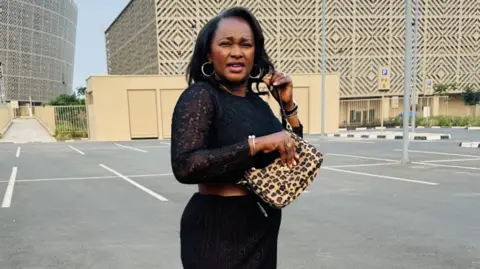 Emertha Uwanyingira Emertha has shoulder length dark hair and is wearing a black dress with a leather belt and lacy full-length sleeves. She has a leopard-print style handbag across her body and is holding it in her right hand. She wears large silver hoop earrings, a silver bracelet and a watch. She is standing in an outdoor car park outside the Petit Stade stadium in Kigali, Rwanda.
