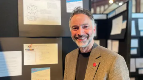 White man with silver hair and a silver blazer smiles in front of a children's design display.