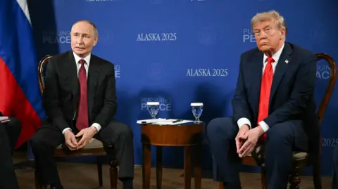 BBC Vladimir Putin and Donlad Trump sit in front of a blue backdrop with the words "Alaska 2025" and "pursuing peace" repeated across it. There is a wooden table between them with two glasses of water. 