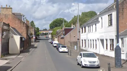 A street with houses on either side. Cars are parked on the right side. A railway bridge is visible in the distance.