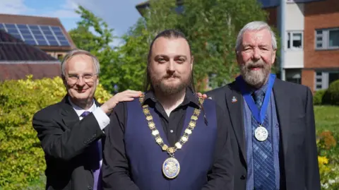 A man with a brown beard and slicked back long brown hair wears a gold chain over a blue waistcoat and a black shirt. He is stood next to two other older men with grey hair. Both men are wearing suits but the man on the right is also wearing a medal with a blue ribbon.