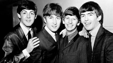The Beatles stood in a group smiling at the camera in 1963