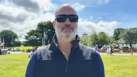 Gareth Watt has very short hair and a grey beard. He's wearing sunglasses and a blue jumper with a gilet. It is sunny and cloudy.
