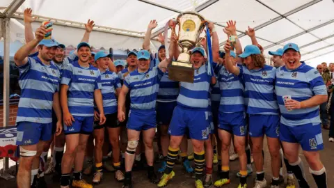 Handout A team photograph of Hospice Barbarians, who wear a striped blue kit with the logo on the top. The team are celebrating together and holding a cup in a marquee.