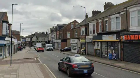 Google Streetview showing a grey taxi driving past a row of two-storey shops. One is a convenience store with the name "Local" written in large yellow letters on a yellow board above the door.