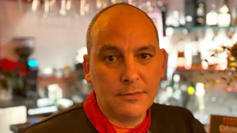 BBC Flavio Serra looks into the camera. He has dark eyes and balding hair. He is wearing a dark chef's outfit with red trims. He is standing at the bar in his restaurant and bottles are visible in the background.