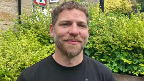 A man called Ross Jackson-Hicks. He has trimmed brown, greying hair. He has a trimmed beard across his lower face. Some of the beard s grey but the rest is brown. He is wearing a black t-shirt and is stood in front of a bush.