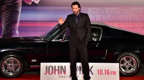 Getty Images Keanu Reeves in a black suit at a John Wick premiere in front of a black sports car