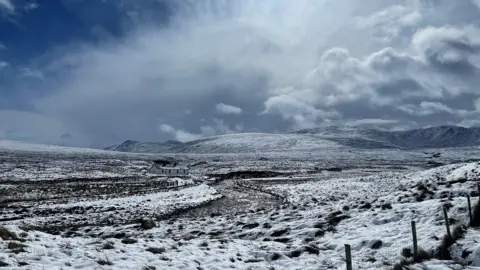Jan Overmeer A wide, snow-covered hillside stretches into the distance beneath a dramatic, cloud-filled sky. A solitary white building sits in the middle of the landscape, with rough ground and fence posts visible in the foreground.