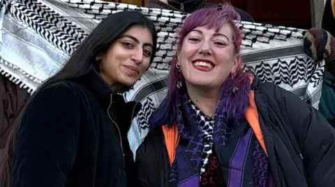 Hiba Ahmed and Ayeshah Behit smile for the camera. Hiba Ahmed has long dark brown hair and is wearing a blue coat with a white floor length, white skirt. Ayeshah Behit has pink/purple hair and is wearing a long black puffa coat, with black trousers and a purple Palestinian-style scarf around her neck.