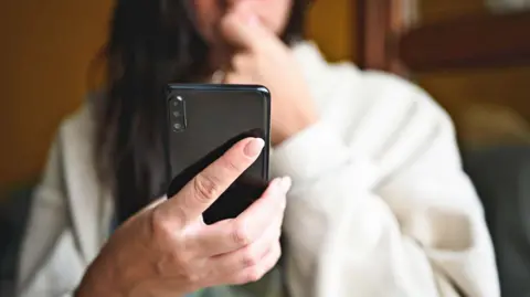 Getty Images A woman with her face out of focus and has long black hair is seen in a white jumper is pictured looking at a black mobile phone.
