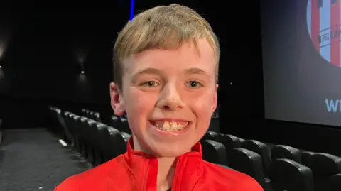 Logan is seen smiling from ear to ear inside a cinema. 
He has short hair and is wearing a Derry City jacket. 
A number of empty seats are seen behind him. 