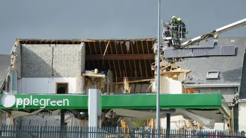 PA Media Two firefighters in an aerial platform appliance above the scene of the explosion at a service station in Creeslough in October 2022. Most of the pitched roof of the front of the building has been blown off. The inside of the wooden rafters are exposed, tiles are missing, solar panels have been displaced and there is a large crack in a grey brick wall which reaches the pitch of the roof.